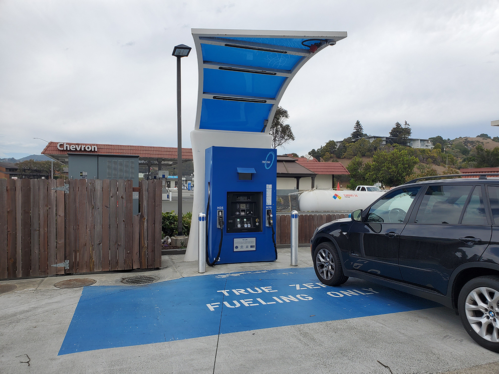 A blue hydrogen fuel cell filling station stands in contrast to the Chevron gas station behind it.