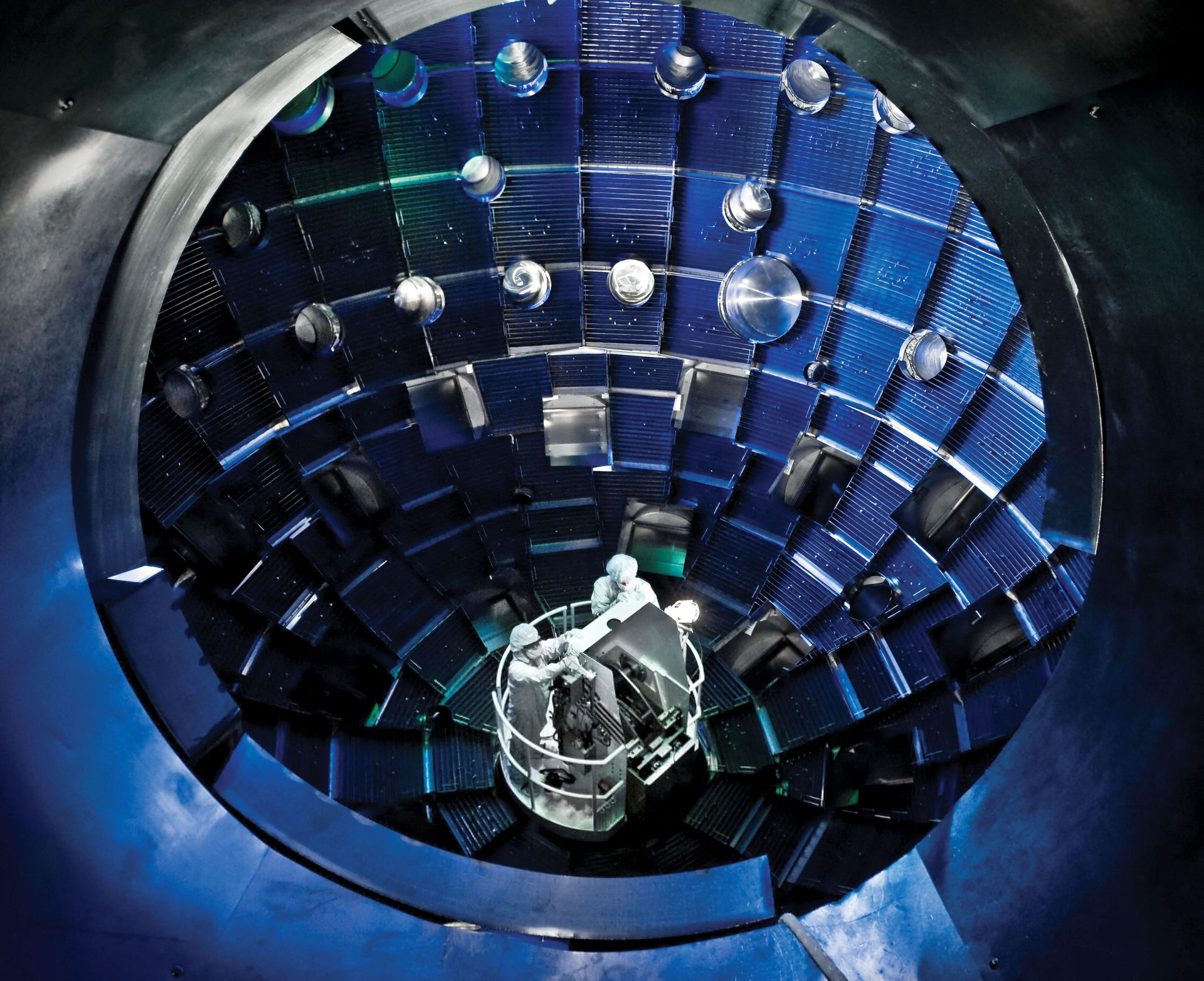 Scientists in white coats operate a device inside of a globular chamber.