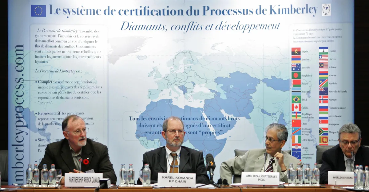 Four men in suits sit at table with microphones for a press conference on the Kimberly Process with a world map with labels in French behind them.