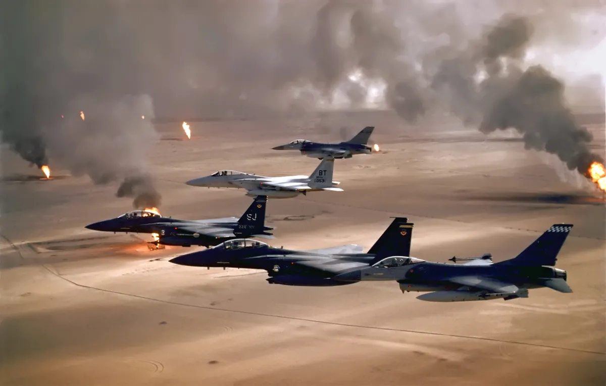 Photo showing U.S. fighter jets flying over a desert landscape spotted with oil well fires spewing plumes of smoke into the air.