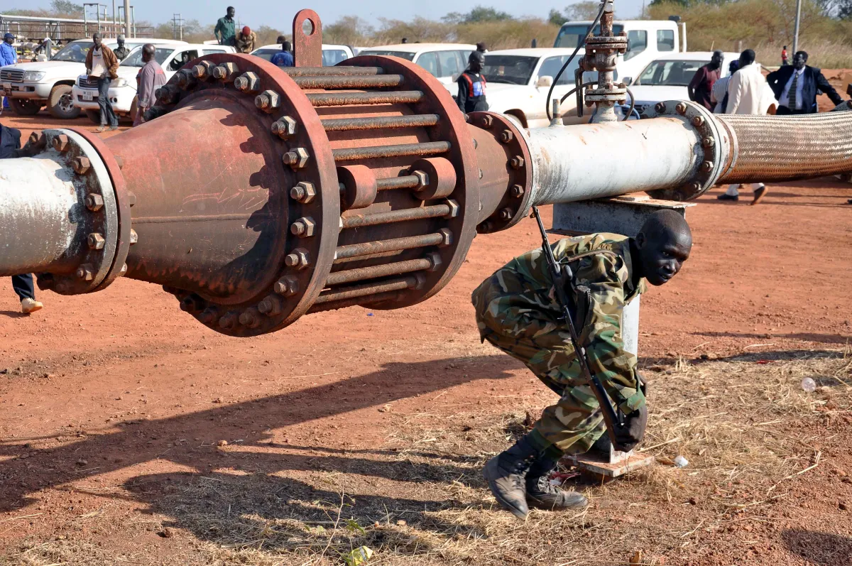 Uniformed man with rifle ducks underneath an oil pipeline.