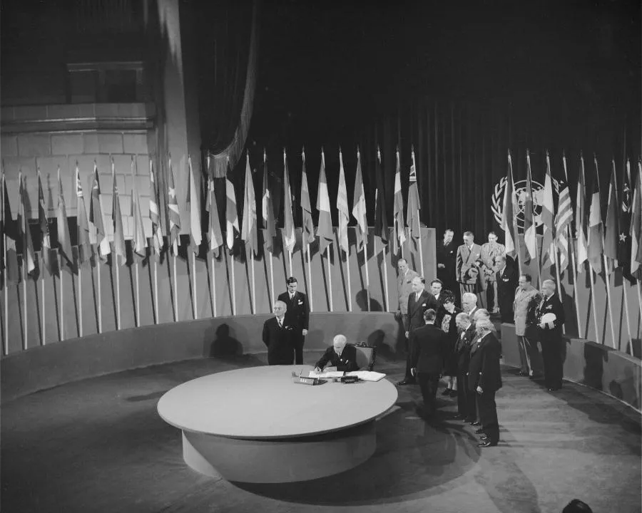 People standing near circular table in front of many different country's flags, participating in the UN charter signing ceremony.