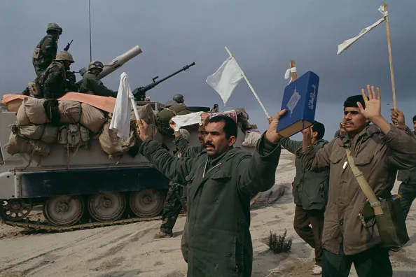 Men hold up their arms in surrender with white flags, as a tank rolls by in the background with armed soldiers on top of it.