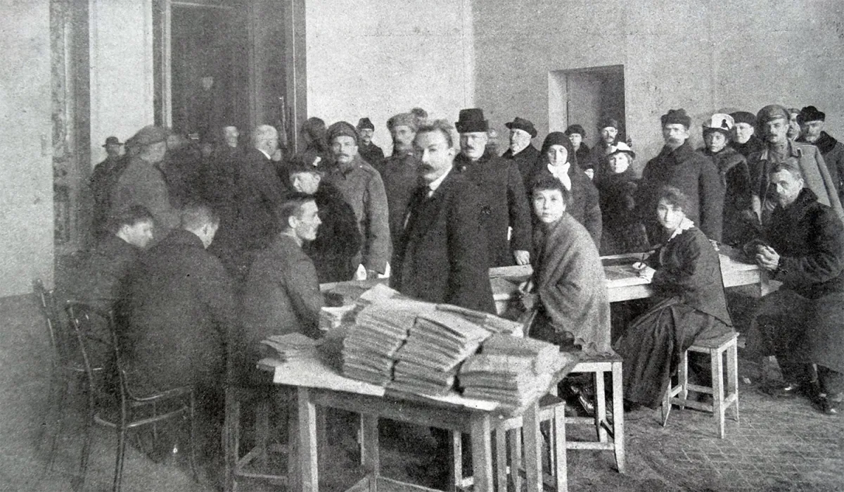 Grainy black and white photograph of people dressed in old-style clothing lined up in room with ballots stacked on tables.
