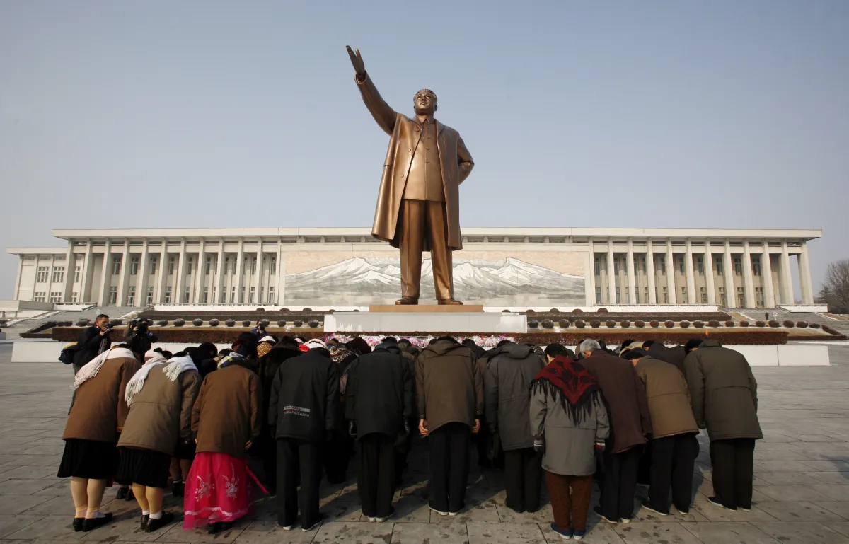 Group of people huddled over bowing down in front of a giant bronze statue of North Korean leader Kim Il Sung holding his arm up.