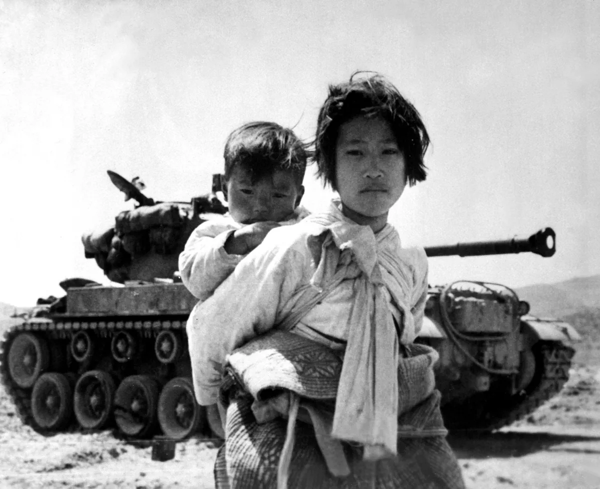 Black and white photo of a young Korean girl holding a toddler on her back, while standing in front of a tank.