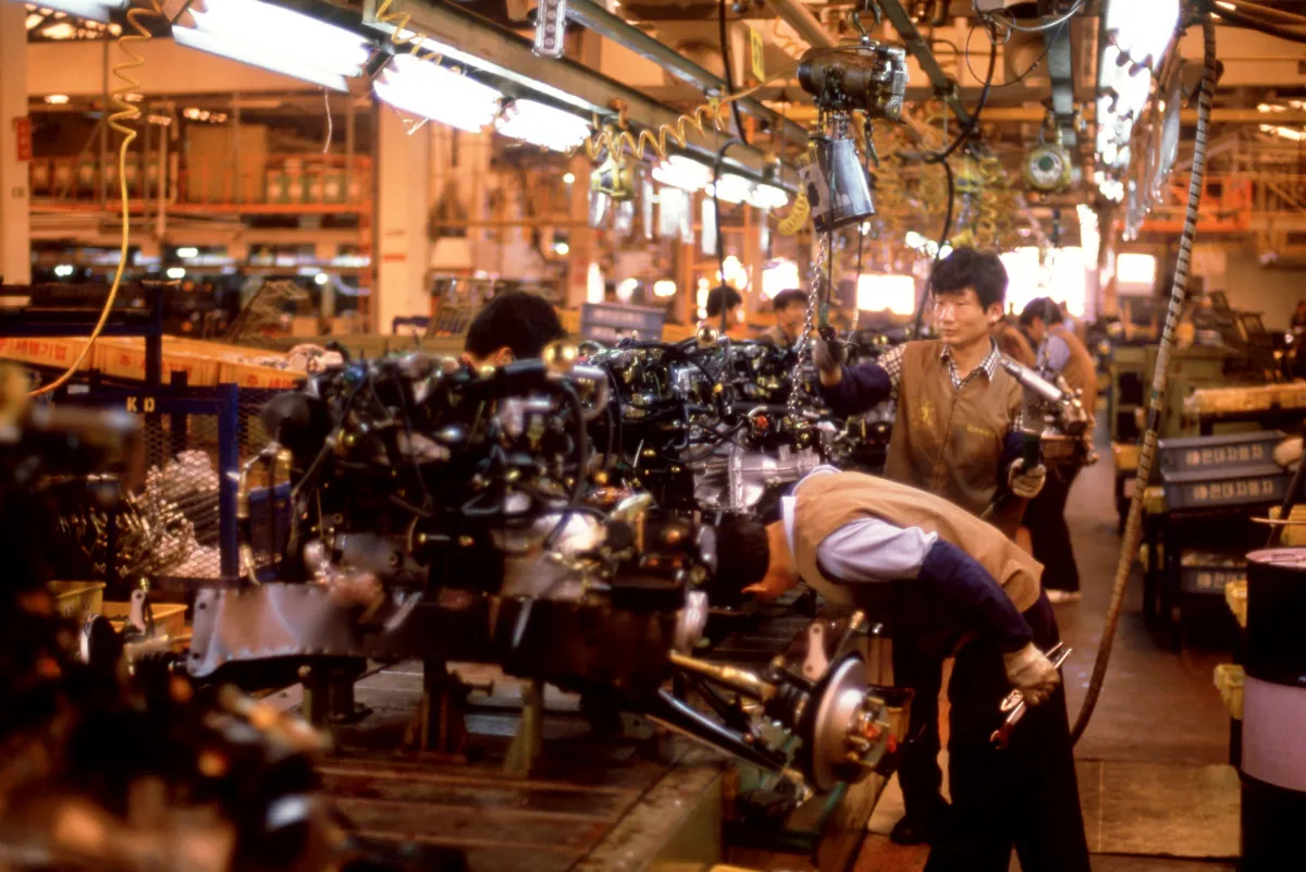 Korean men wearing brown uniform vests while working on an assembly line with many car engines.